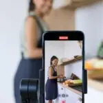 Woman vlogging her cooking tutorial in a kitchen using smartphone on a tripod.